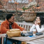 A man and a woman sitting across from each other at an outdoor wooden table, smiling and talking, with notebooks, a coffee cup, and a backpack on the table.