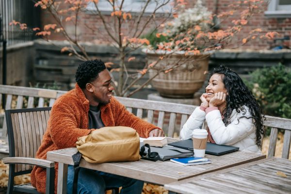 A man and a woman sitting across from each other at an outdoor wooden table, smiling and talking, with notebooks, a coffee cup, and a backpack on the table.