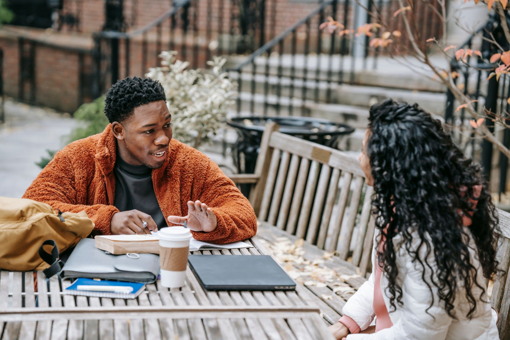 A man gesturing as he talks to a woman seated across from him at an outdoor table with notebooks, a coffee cup, and a backpack.