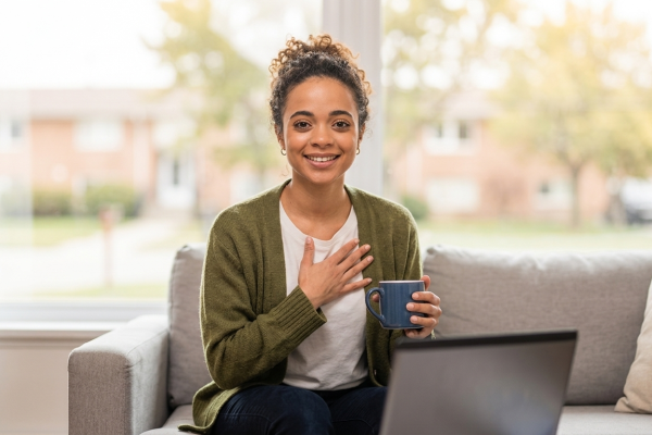 A smiling woman sitting comfortably on her couch holding a mug during an online counselling appointment for anxiety